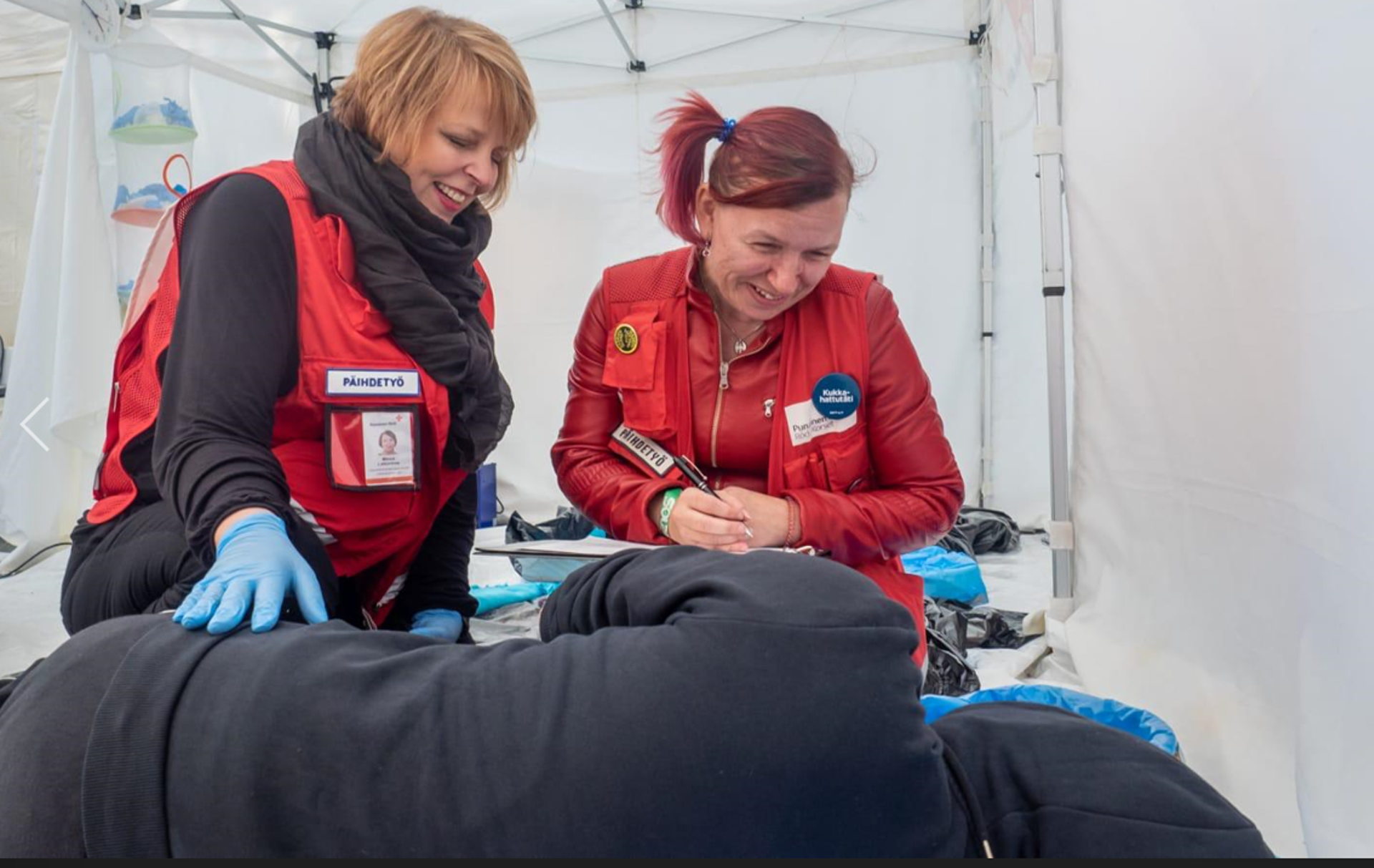 In the Red Cross first care tent, two volunteers wearing red vests are helping a person lying on an examination table.
