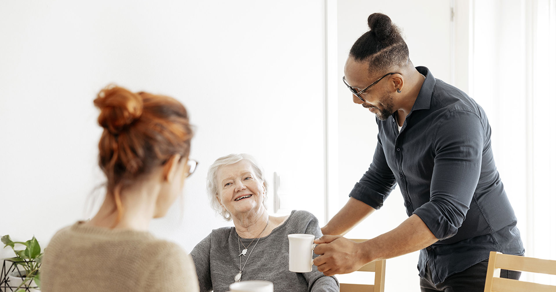 Two people sitting. A third person hands them a small jug.