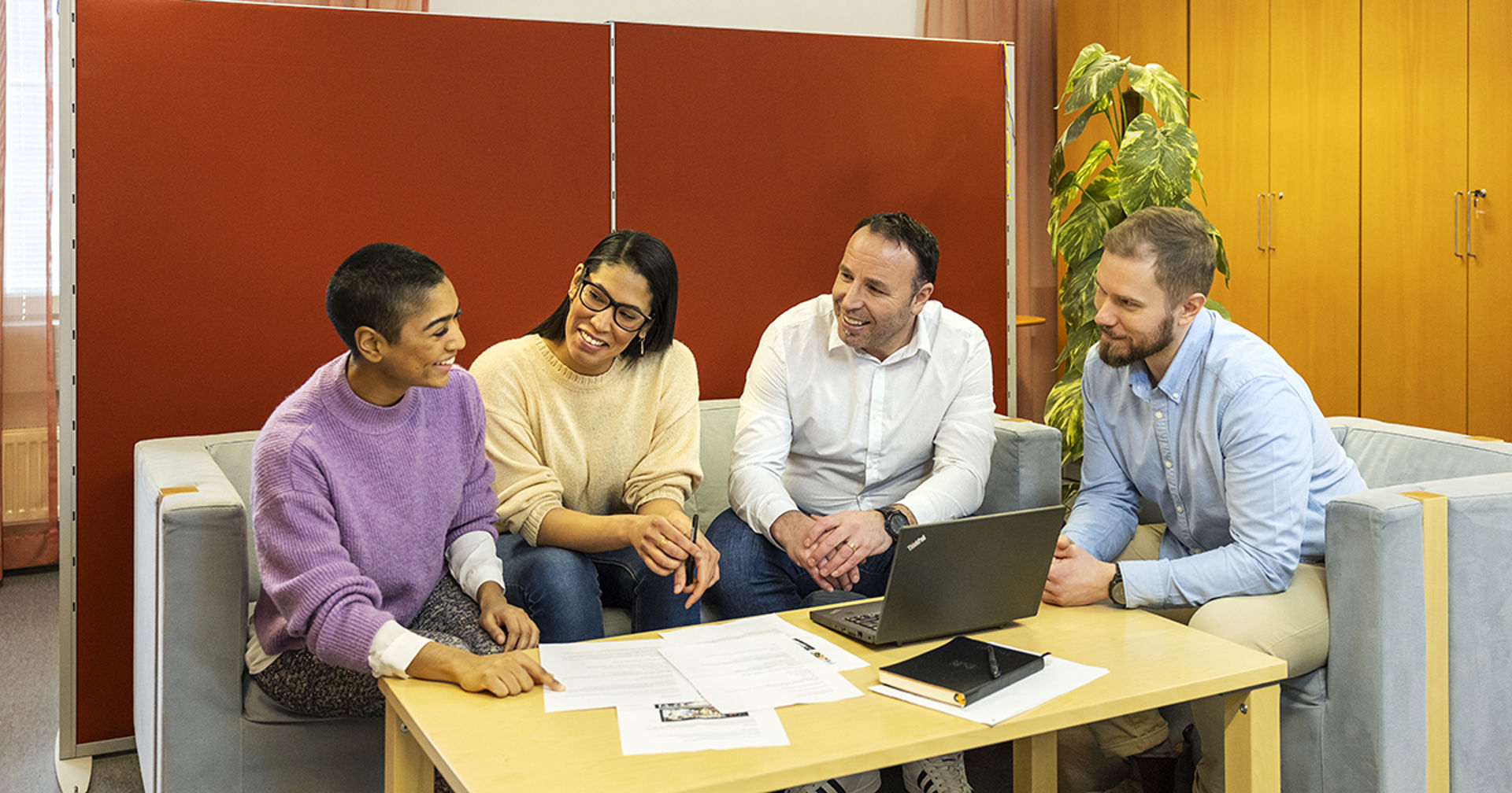 Four people sitting around a table and smiling. There is a laptop, some papers and a book on the table.
