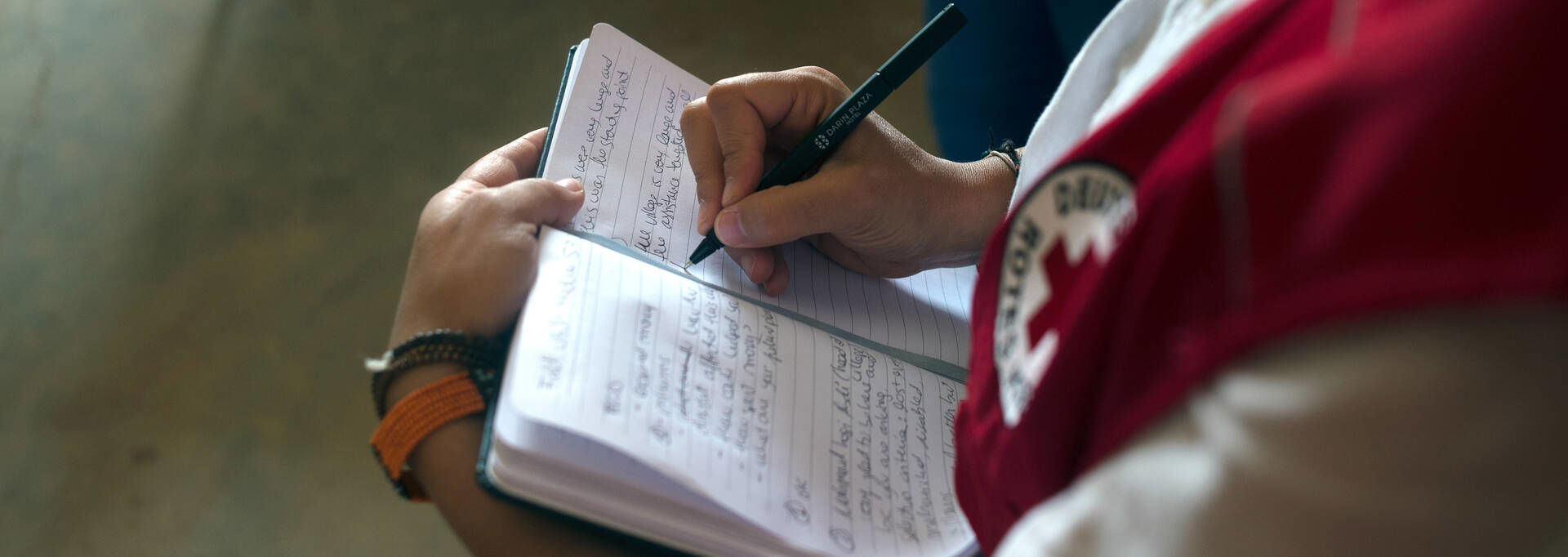 A person wearing a Red Cross vest is writing down to a notebook.