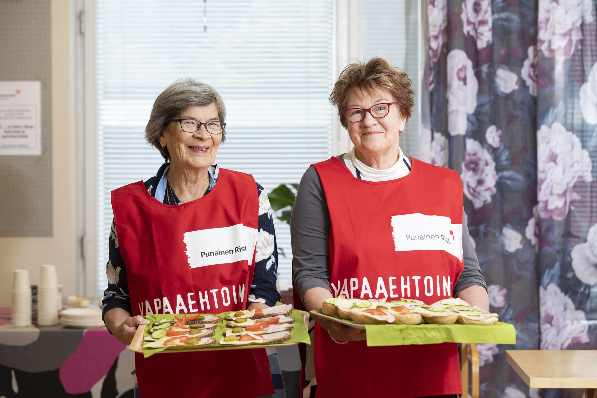 Two volunteers are smiling at the camera and holding trays with sandwiches.