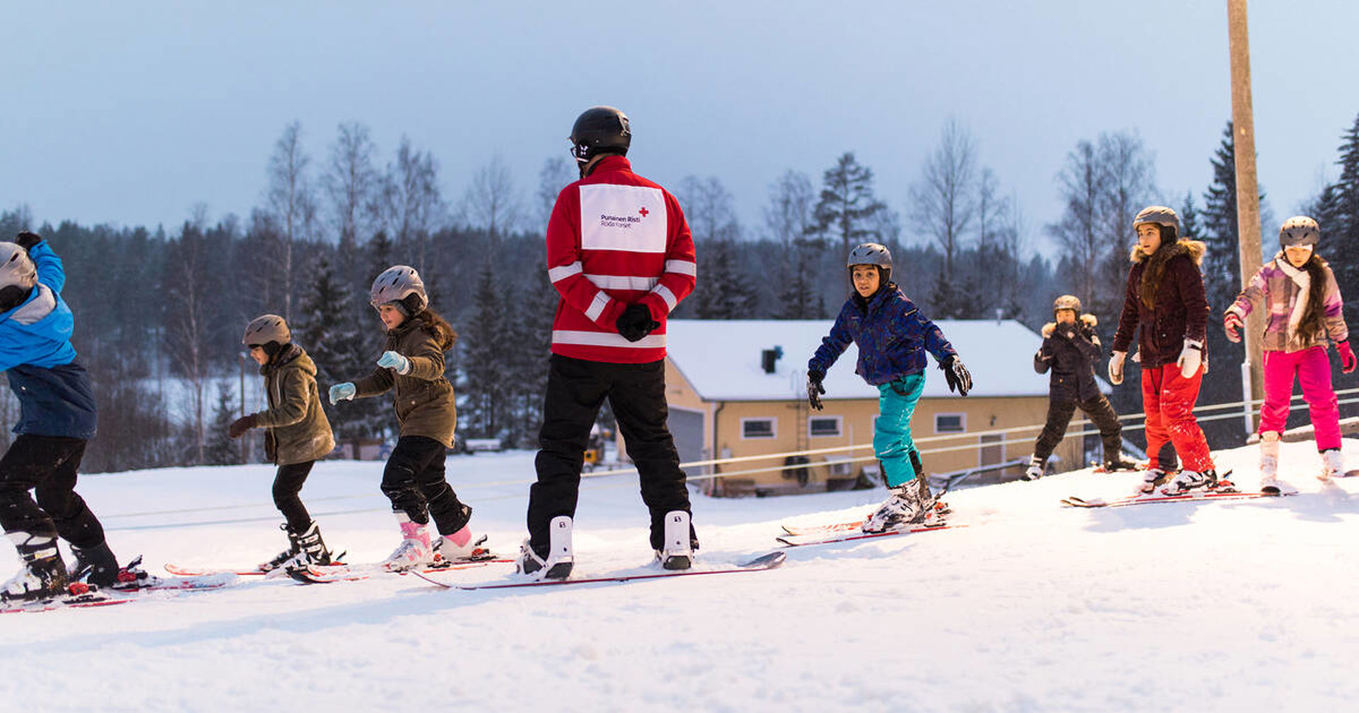 An instructor wearing a Red Cross jacket teaching a group of children how to ski.