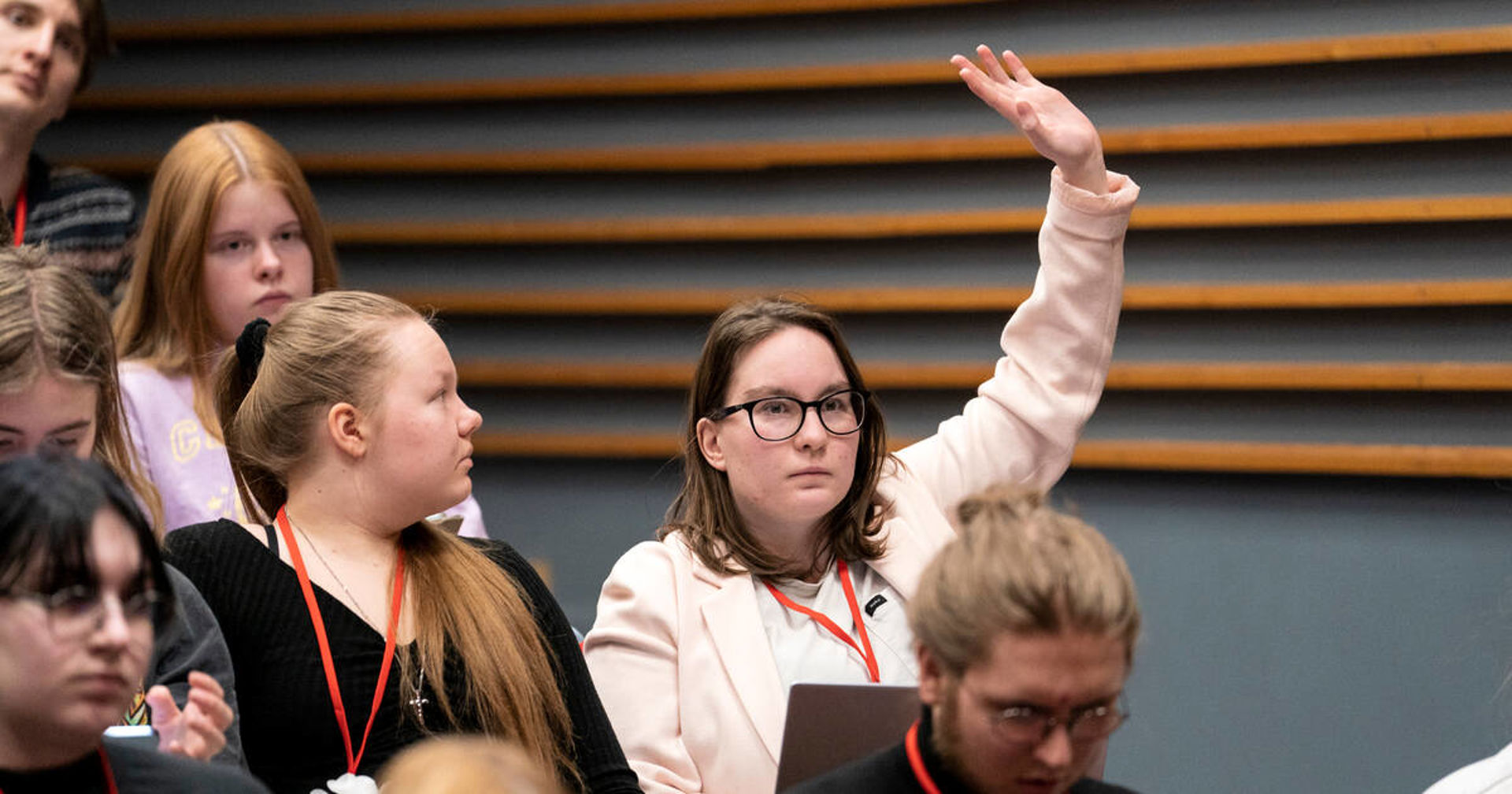 The image shows young people in a meeting. One of the young people is raising their hand to request a turn to speak.