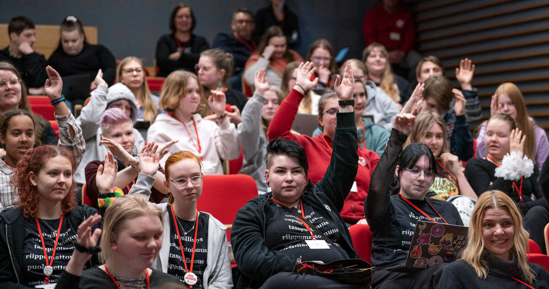 In the picture, there are several young people at a meeting. They are all raising their hands, requesting a turn to speak.