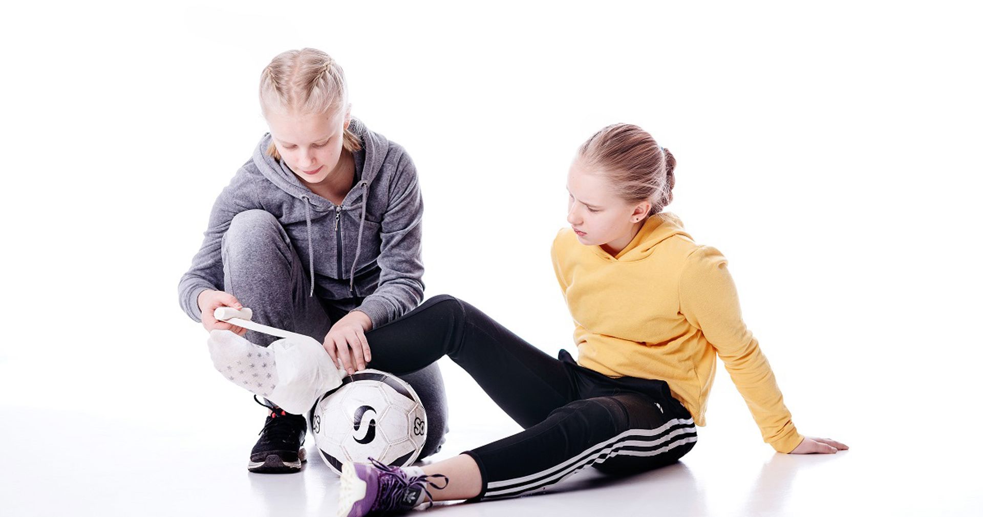 Two kids sit on the floor with a football. On is tying a bandage around the others ankle.
