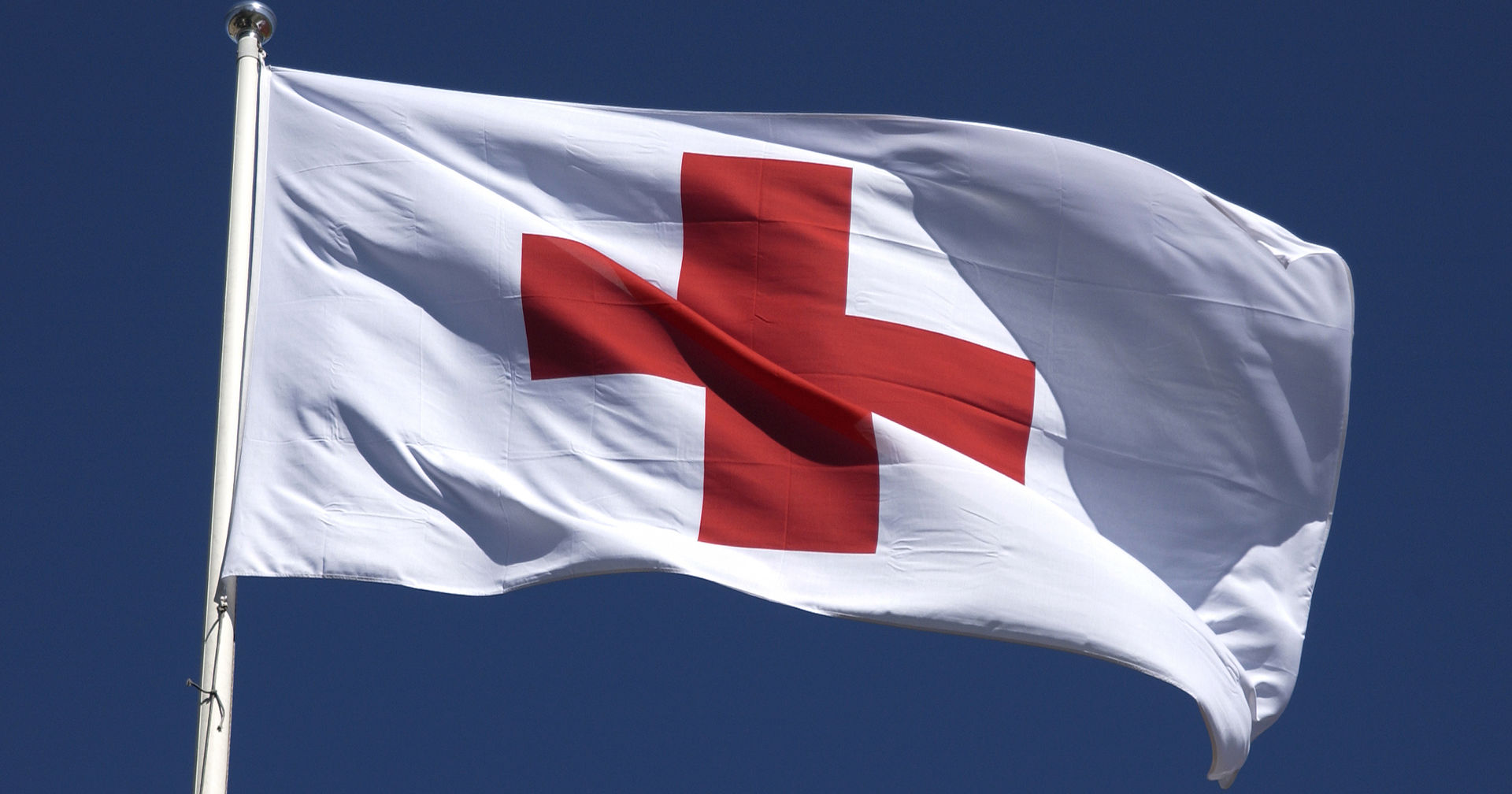 The Red Cross flag flying on a flagpole against a blue sky.