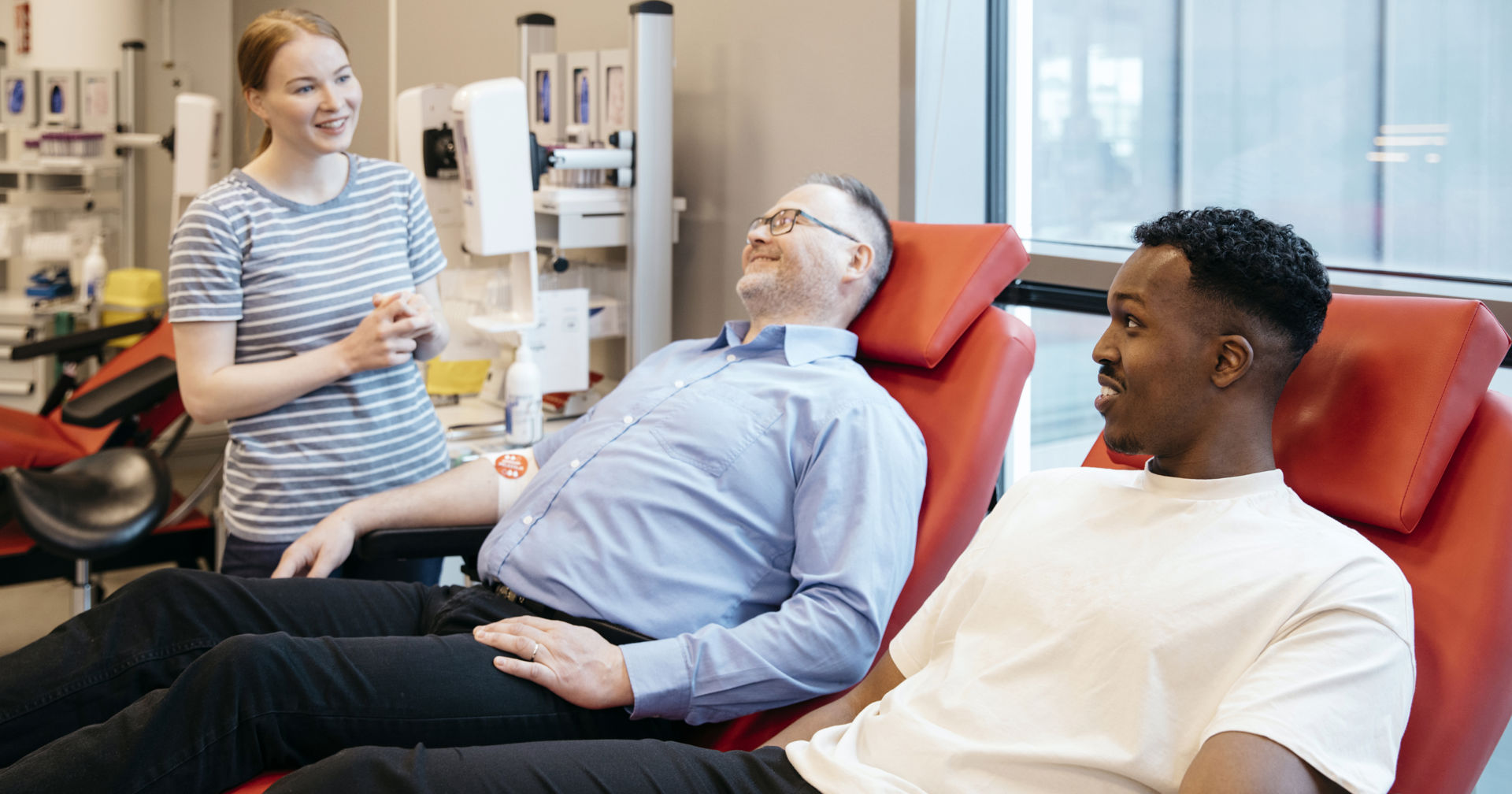 Two people are smiling at a nurse after blood donation. They have bandages and a Blood Service sticker on their arms.