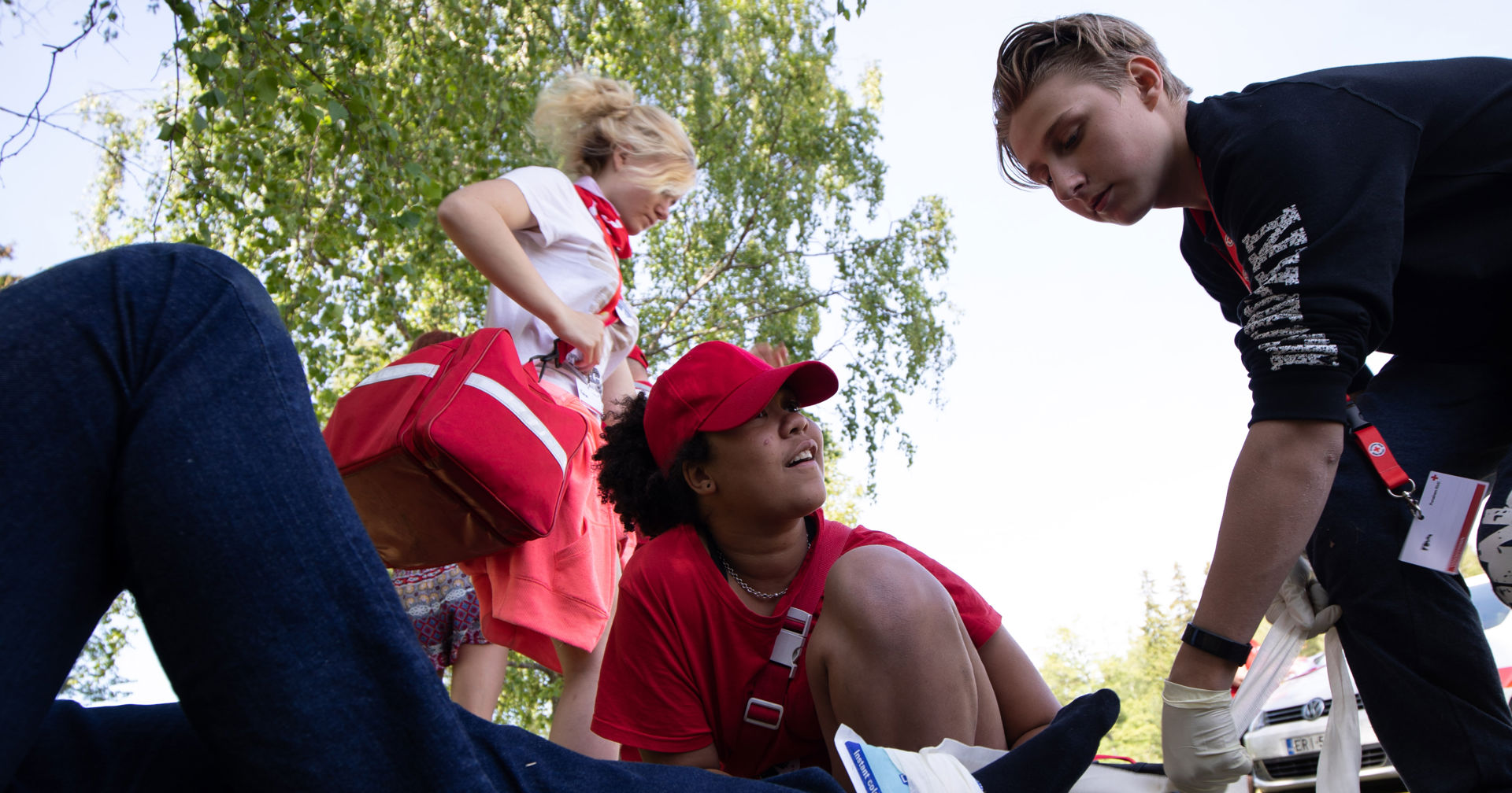 Two young people in Red Cross shirts are tying an ice pack to someone's ankle. In the background, a third young person is carrying a red first-aid kit