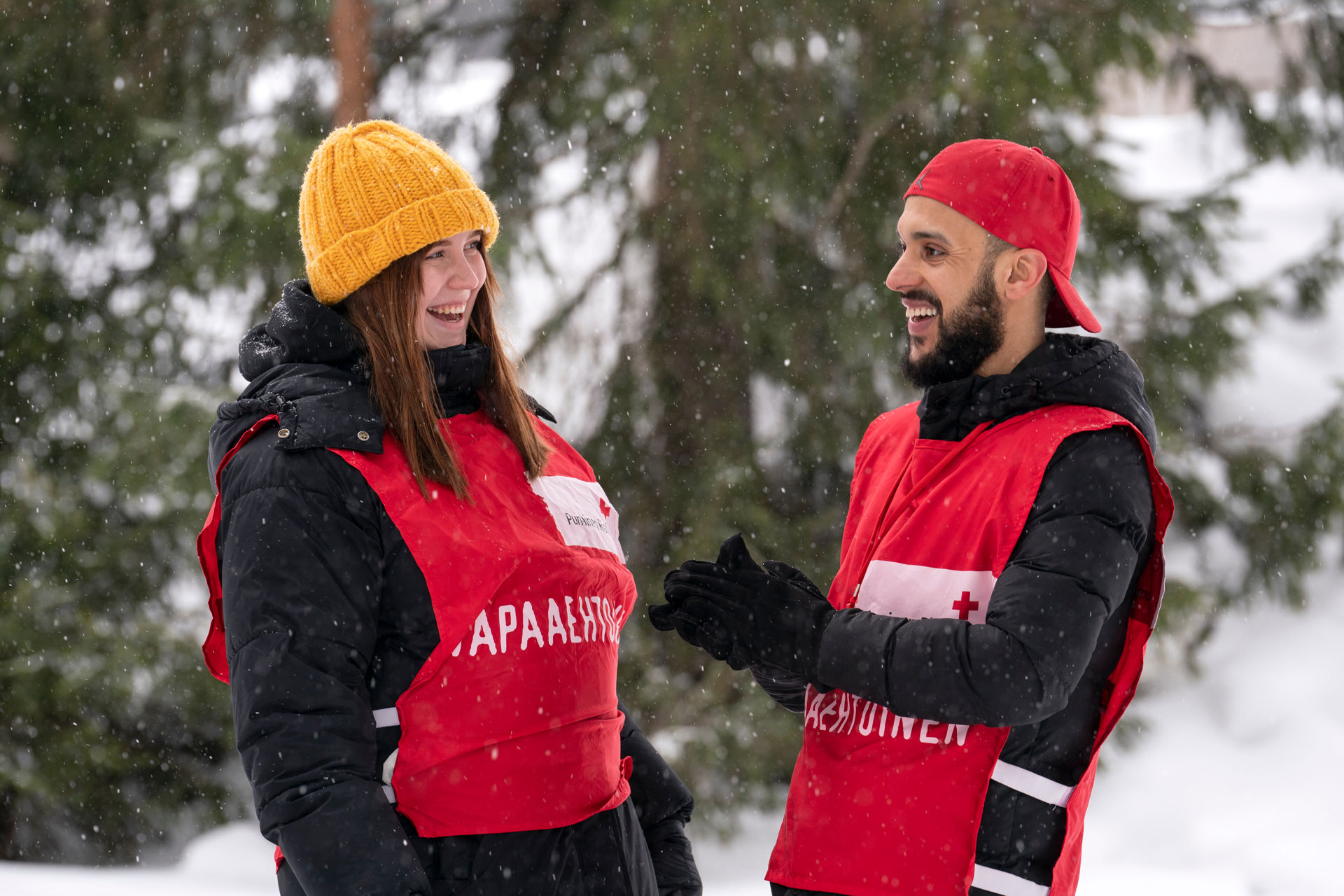 Two Finnish Red Cross volunteers standing outdoors in a snowy forest in winter clothes and red vests with the Red Cross emblem.