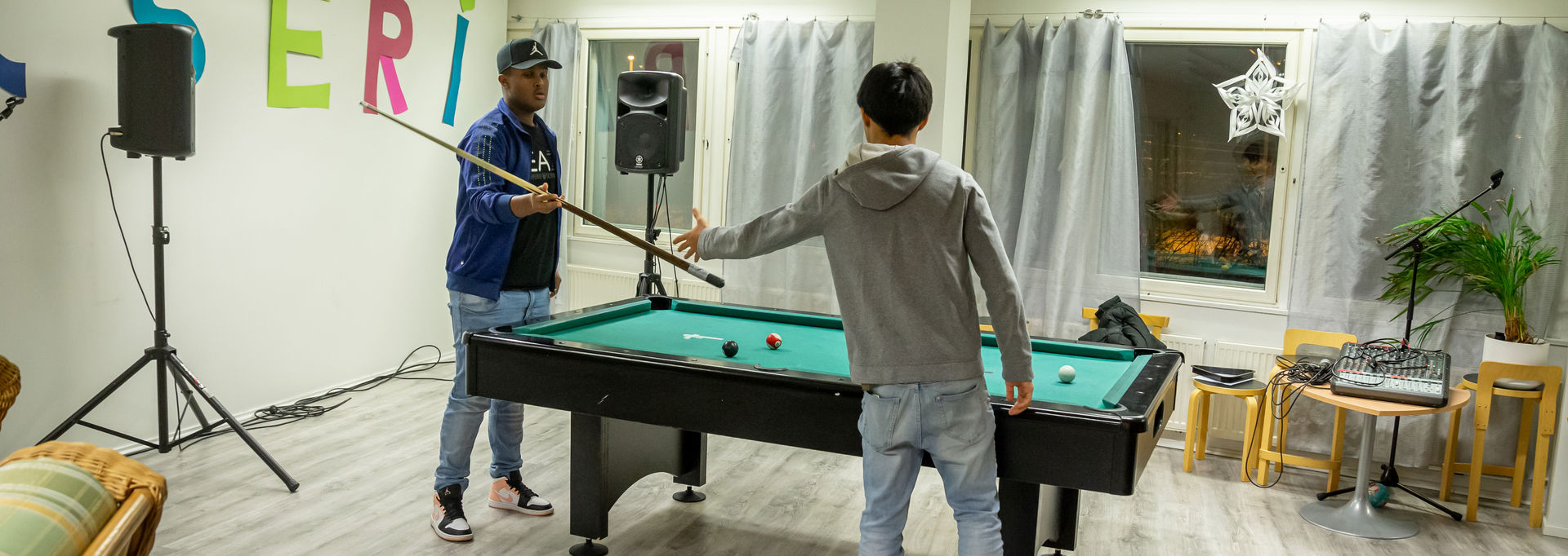 A young person passing a pool cue to another young person. They are standing by a pool table.