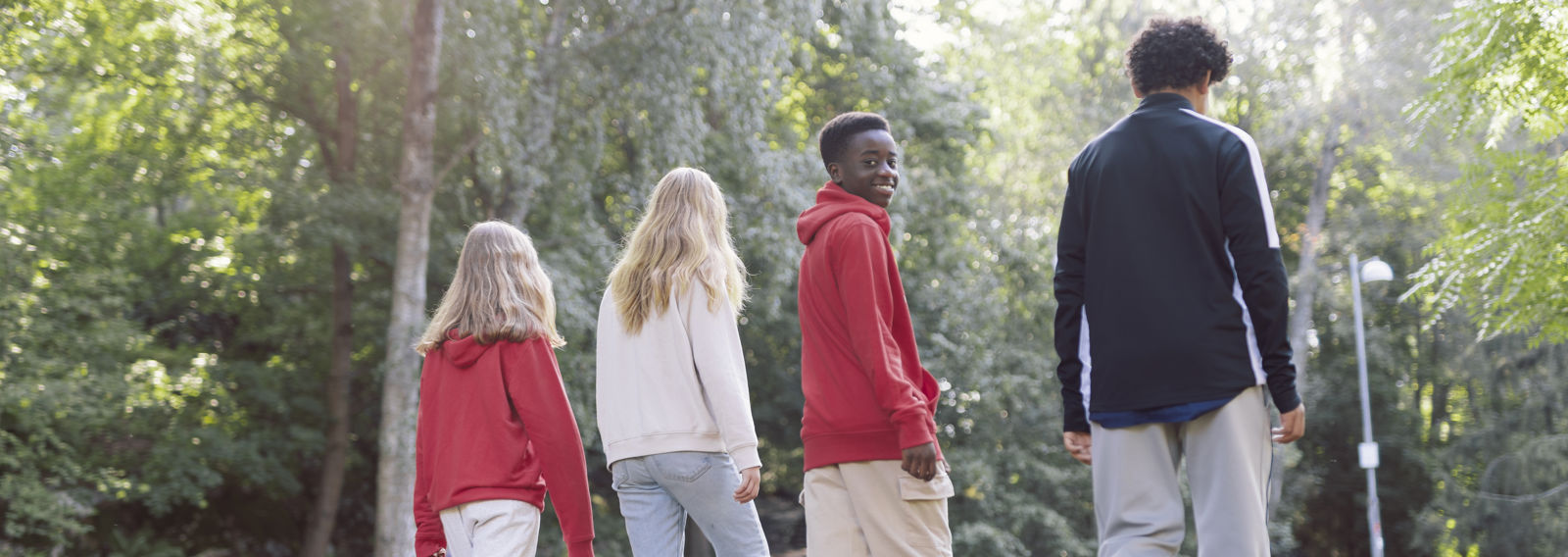 Four young people are walking in a park with their backs to the camera. One of them has turned their head and is smiling at the camera.