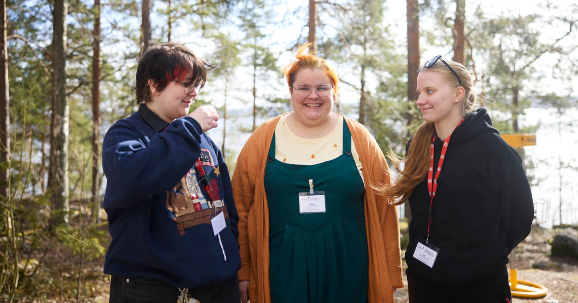 Three young Red Cross volunteers are standing side by side outdoors and chatting cheerfully. The person in the middle of the image is looking towards the camera.