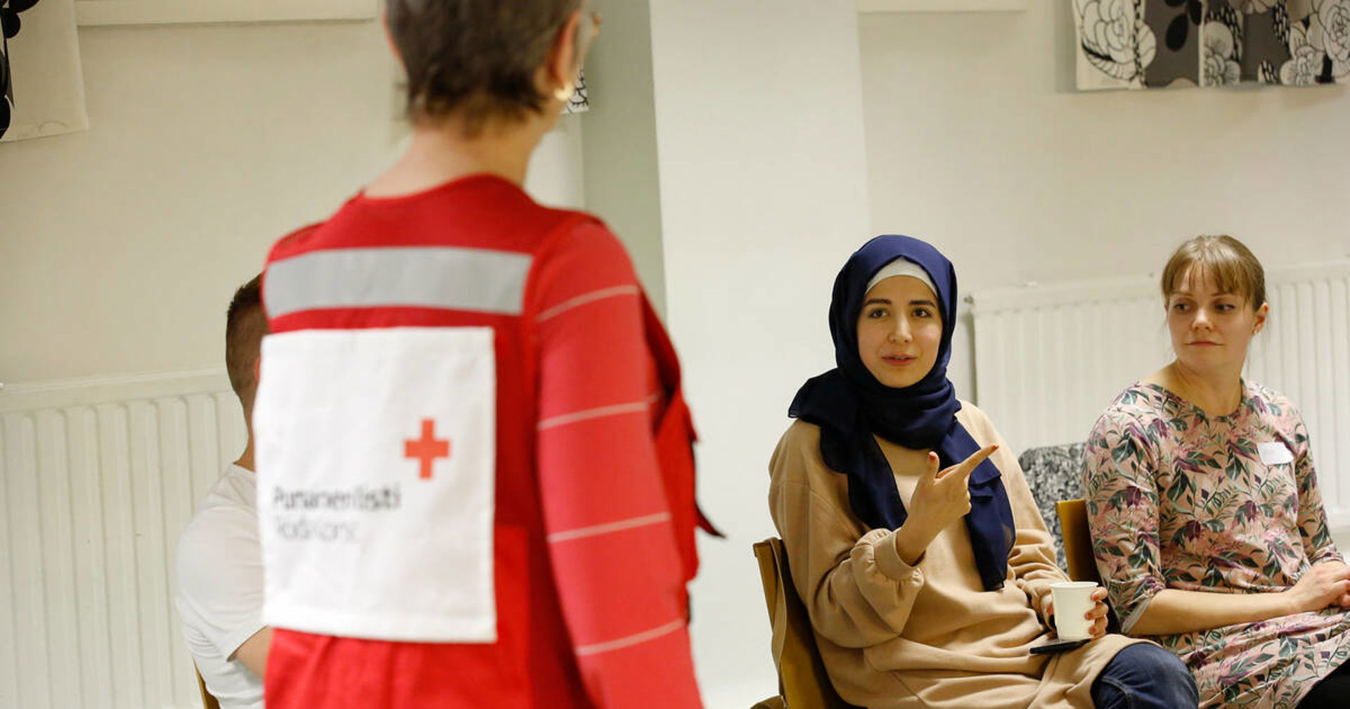In the photo, with their back to the camera, is a volunteer trainer wearing a red volunteer vest. Additionally, two young volunteers are sitting in the photo, facing the camera.