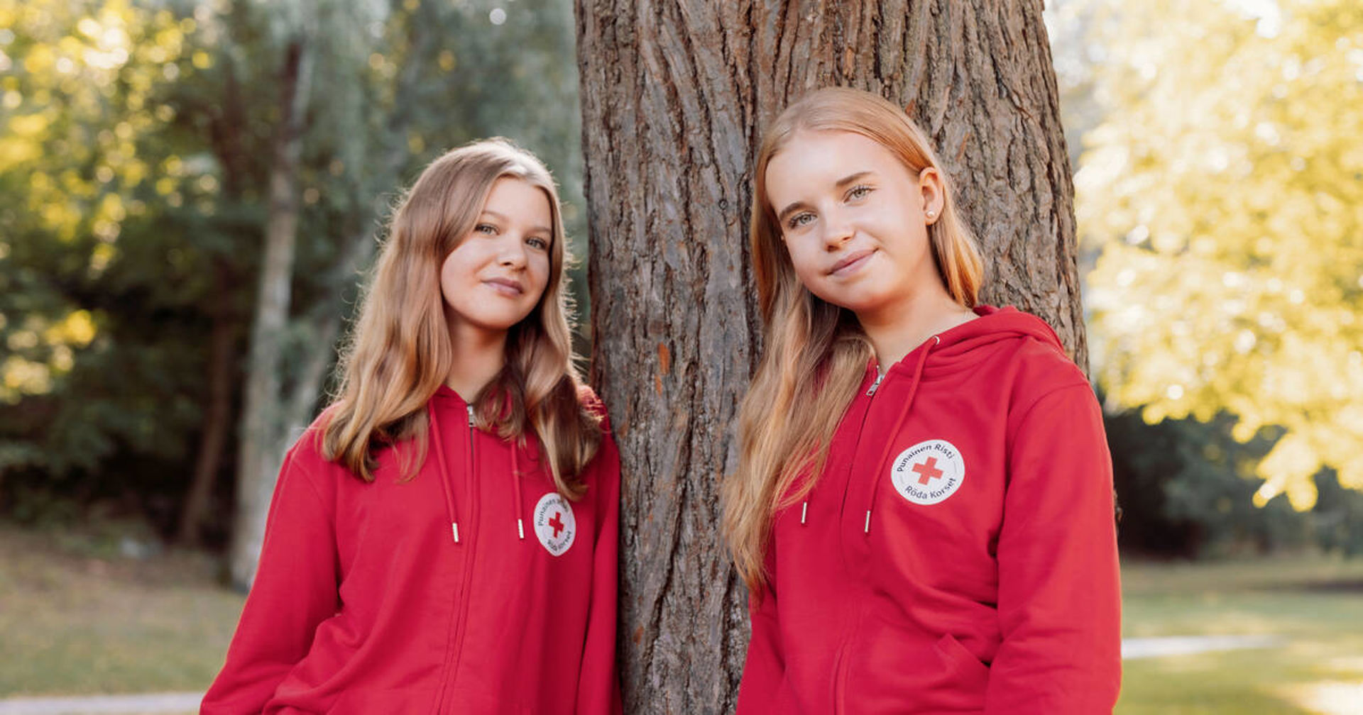 Two young people are leaning against a tree outdoors. They are wearing red Red Cross hoodies.