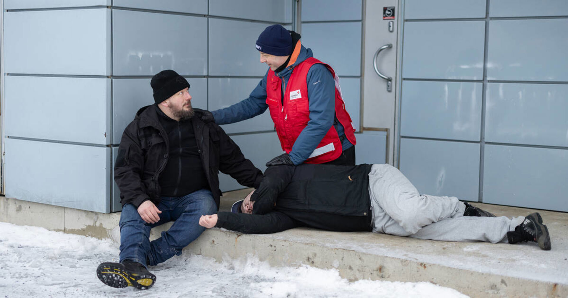A Red Cross volunteer in a red vest helps two people outside a building in winter conditions. One person is sitting on a step and the other is lying on their side on the ground.
