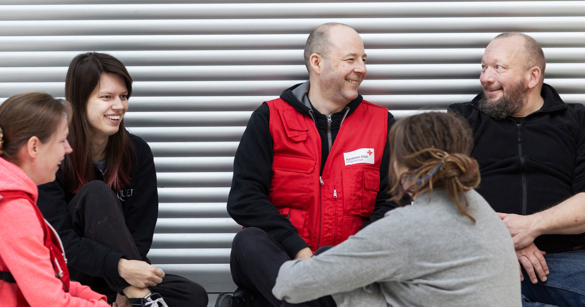 A group of people are sitting and discussing. One person is wearing a red vest from the Red Cross.