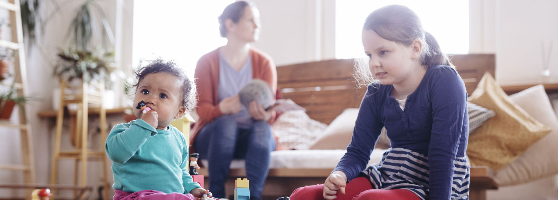 Two children are playing with toys in the foreground, and a little child has put a toy in their mouth. In the background, a parent is reading a magazine.