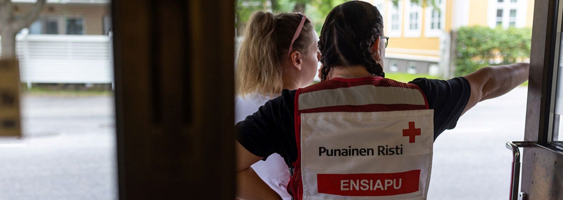 A volunteer working at the Red Cross first aid duty service stands in a doorway and points the way for another person. The text ”Finnish Red Cross First Aid” is visible on the volunteer’s vest.