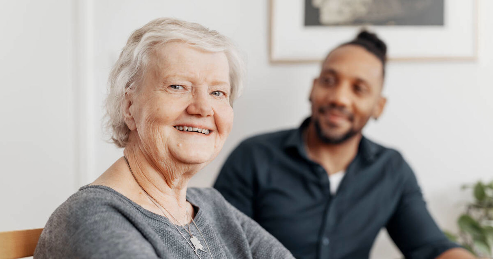 An older person looks at the camera. In the background, there is a person looking at them with a smile.